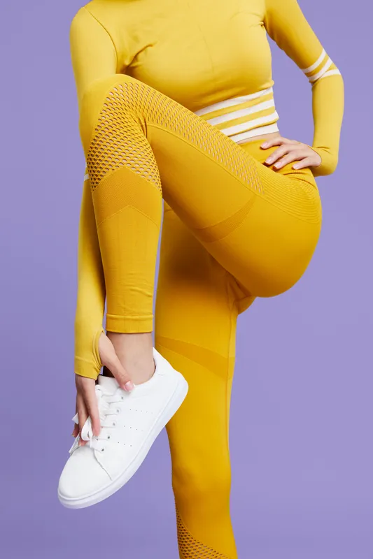Fit woman sport fitness dressed in yellow activewear adjusts white sneaker, preparing for workout on vibrant purple background, showcasing strength and flexibility.