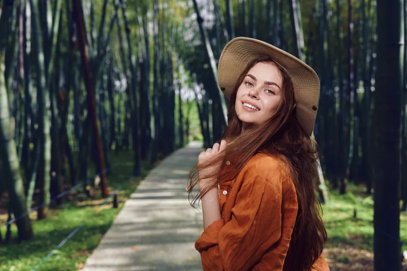 Woman in straw hat smiling on a bamboo path, portrait of longhair female outdoors in orange jacket. Walking on a forest walkway with sunlight, nature backdrop and relaxed travel mood.