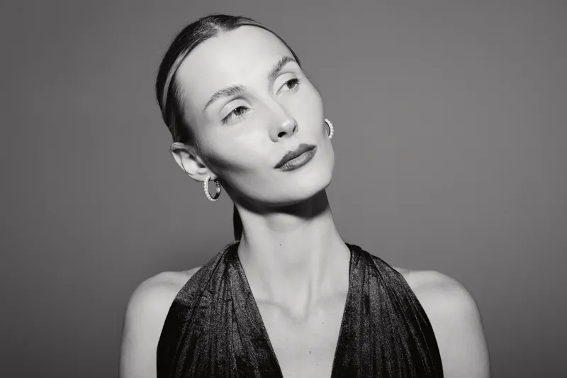 Portrait of a fair skinned Caucasian woman with slicked back hair wearing hoop earrings and black dress on gray background.