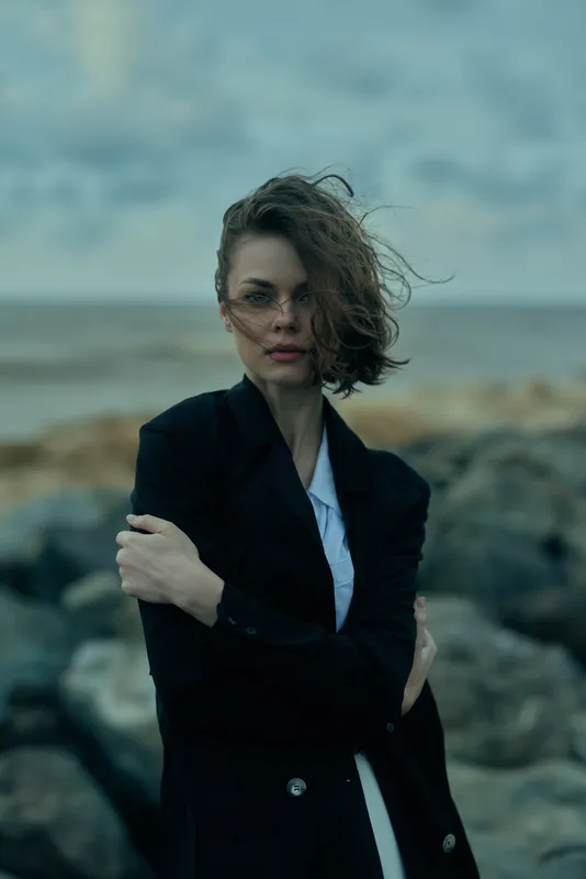 Woman in a tailored dark coat stands on a windswept rocky coastline, hair tousled, moody atmosphere, fashion forward look, strong stance against the sea wind