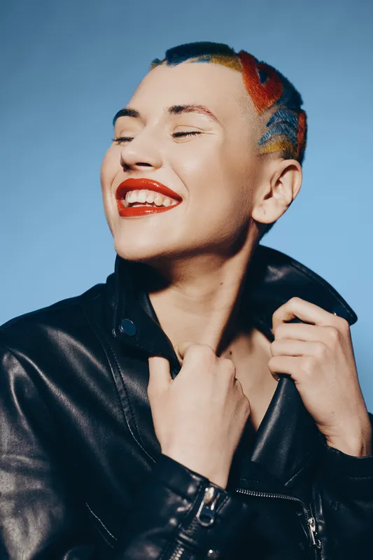 Smiling woman with colorful short hair wearing black leather jacket, expressing joy and confidence against plain blue background in studio lighting.
