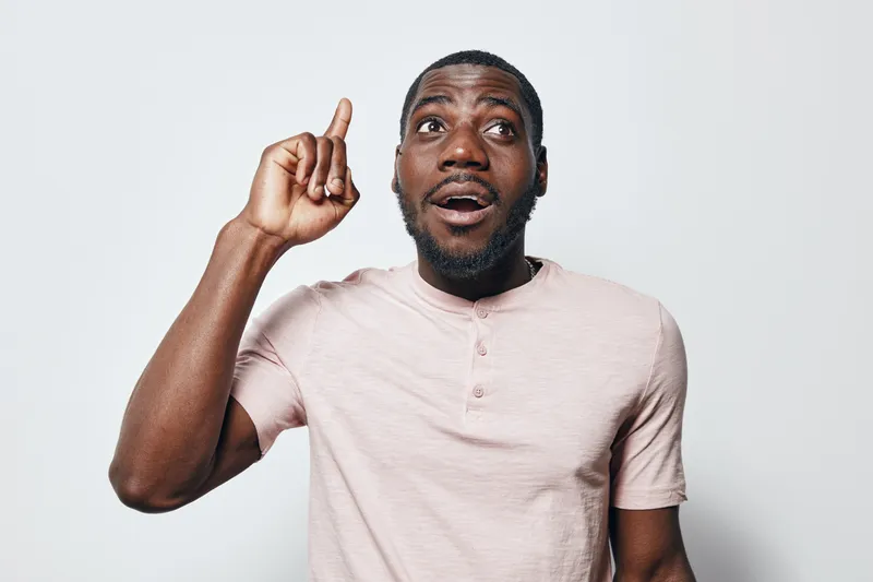 African American man with dark skin wearing light pink shirt pointing up with finger and looking surprised or thoughtful on white background. Expression, gesture and casual style.