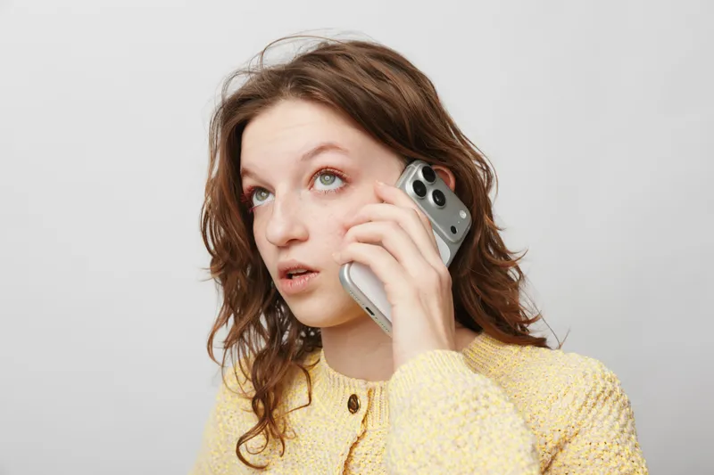 Young woman with curly hair wearing yellow sweater talks on smartphone looking up with thoughtful expression. Concept of communication, technology, and casual lifestyle.