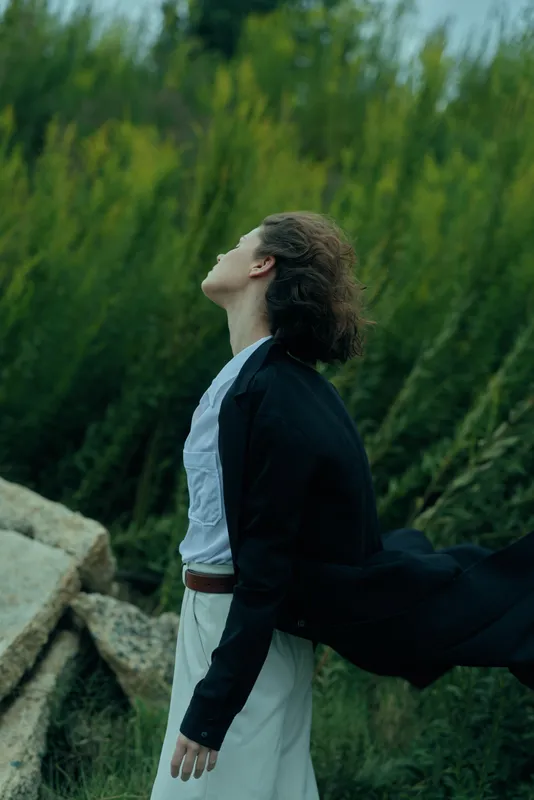 Woman in a black coat and white dress stands in a windy field, gazing skyward as a cloak flows behind, conveying movement, freedom and calm against green scenery