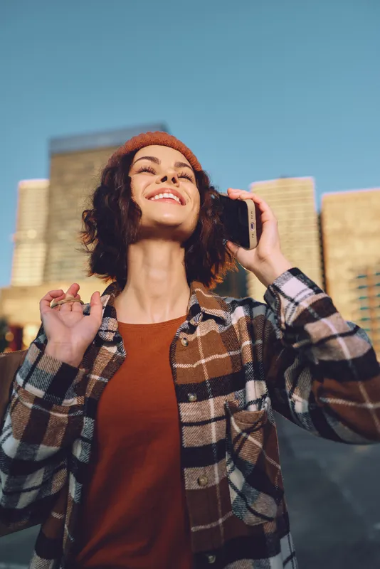 Woman on phone smiling in urban street during golden hour, candid lifestyle portrait with authenticity and emotional storytelling, beanie and flannel jacket, joyful mindful living vibe.