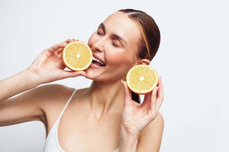 Woman holding orange slices, citrus freshness, smiling with relaxed eyes, showcasing healthy vitamin intake, beauty and skincare vibe, clean studio background, soft light