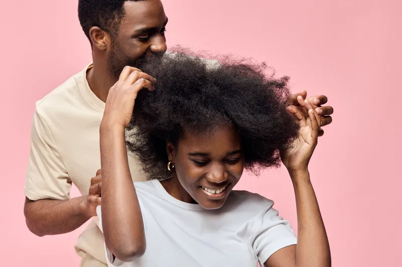 Happy young couple enjoying playful moments while laughing and embracing, showcasing their natural hairstyles against a soft pink background, ideal for relationship concepts