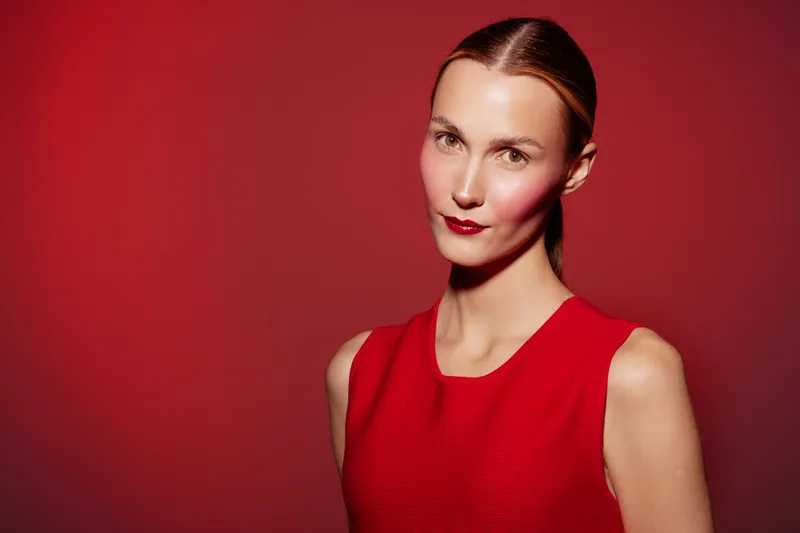 Portrait of a confident woman in a red dress standing against a red background. The image highlights elegance, fashion, and beauty with soft lighting and a serious expression.