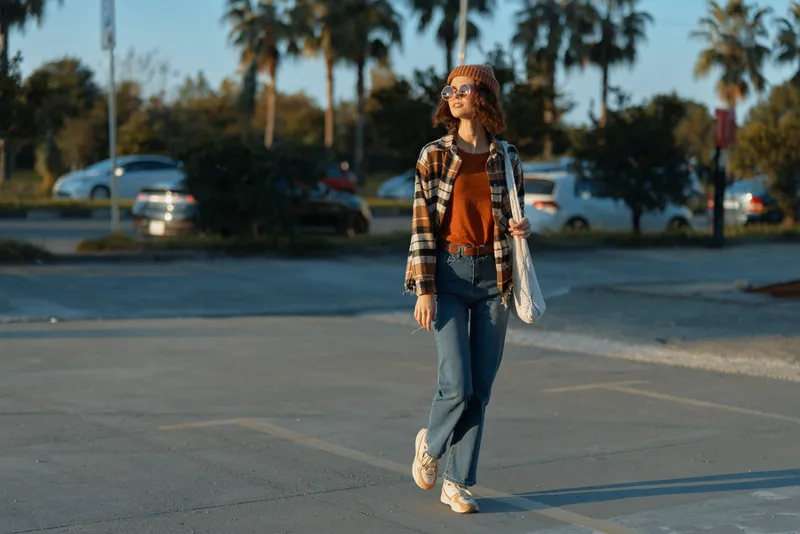 Woman walking at sunset in casual urban fashion, sunglasses and beanie, carrying a tote in a parking lot. Candid lifestyle portrait with authenticity, golden hour glow and emotional storytelling.