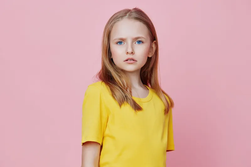 Young girl with long blonde hair wearing a yellow t shirt against a pink background, embodying innocence and youthfulness in a vibrant setting