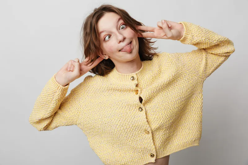 Playful young woman in yellow sweater makes funny face and peace signs with fingers. Casual studio portrait on light background. Concept of youth, humor, and carefree attitude.