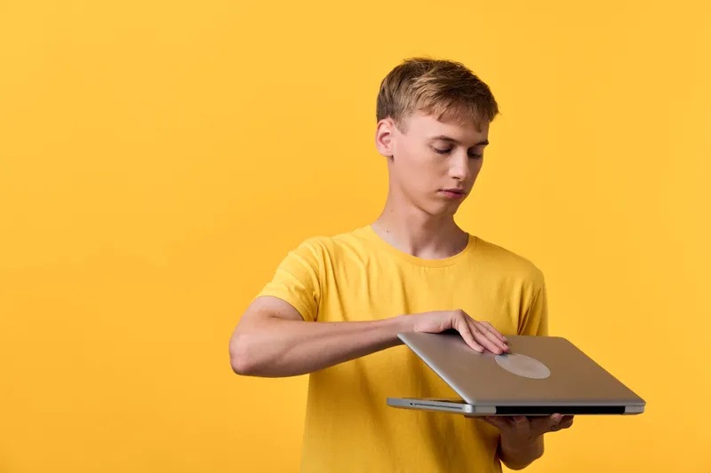 Young man in a yellow shirt opens a laptop lid toward the camera, showcasing modern technology in a bright studio, focused expression, clean composition, daily device for learning and creativity