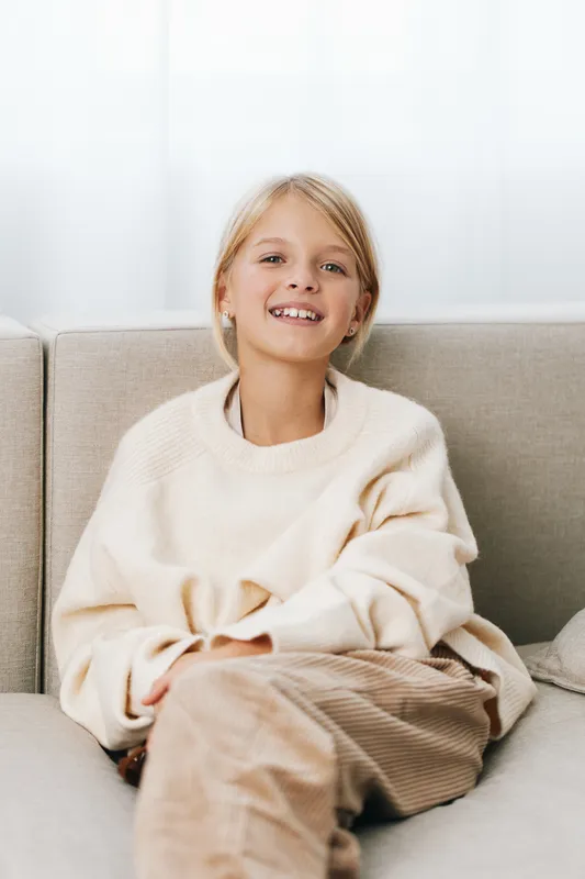 Authentic portrait of a young girl relaxing on a couch, wearing a cozy sweater, embodying a playful school lifestyle moment framed by natural light and calm mood.