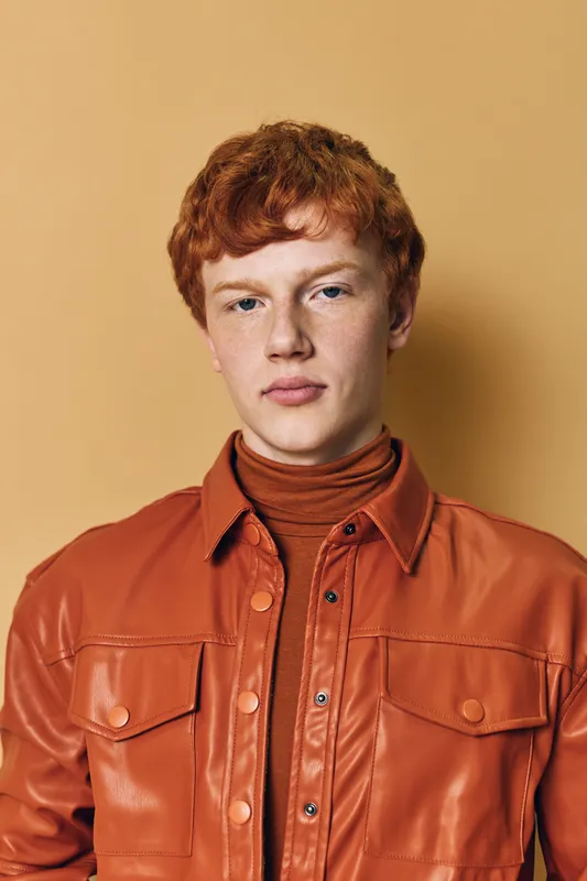 Young man with red hair wearing orange leather jacket and turtleneck posing against beige background in studio portrait with serious expression and direct look.