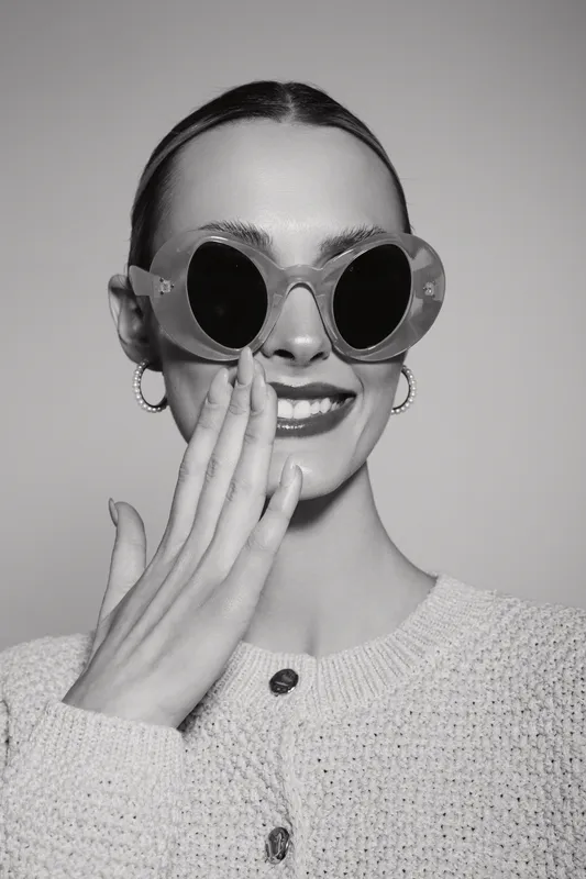 Smiling young caucasian woman with light skin wearing round sunglasses and knitted sweater covers mouth with hand, studio portrait in black and white.