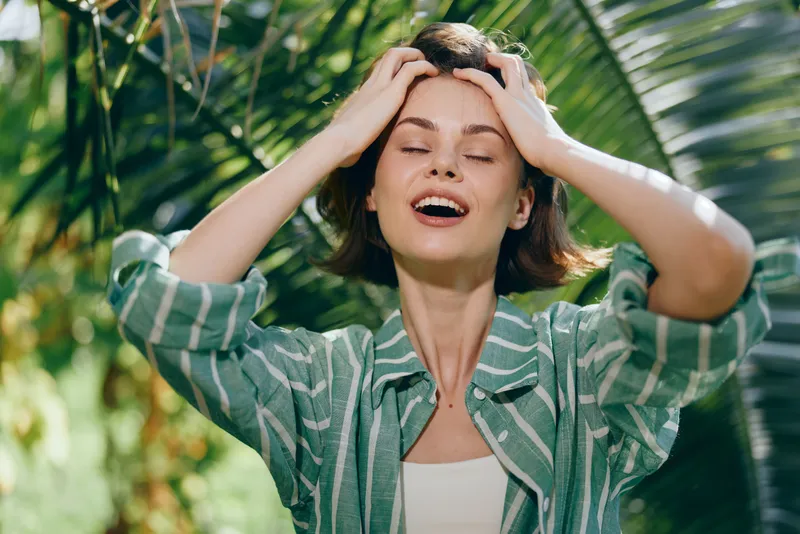 Joyful woman outdoors, eyes closed, hands in her hair, smiling beneath palm leaves, sunlit and carefree.