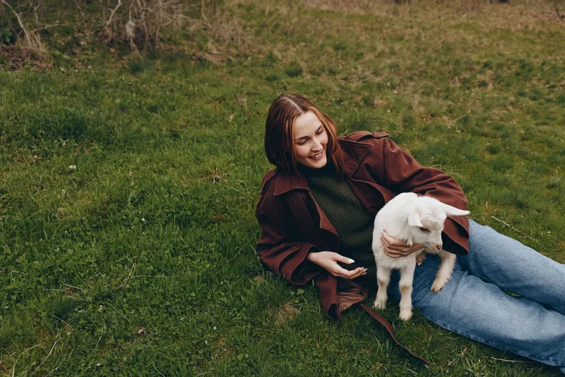 Relaxed woman lying on grass with a playful goat kid, enjoying outdoor life and calm countryside vibes. Casual clothing, soft daylight, conveying companionship, wellness, mindfulness, and gentle