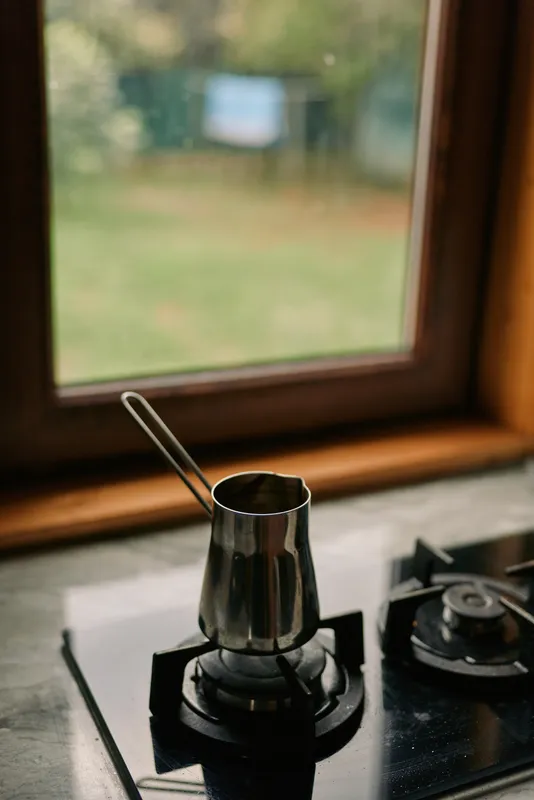 Coffee cezve on gas stove by kitchen window morning scene with metal pot on burner steam rising cozy countertop view of blurred garden light and simple home brewing ritual