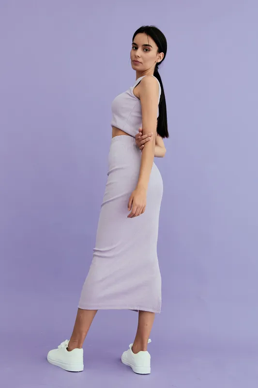 Young woman in casual striped two-piece outfit stands confidently against a pastel purple background wearing white sneakers and minimal makeup.