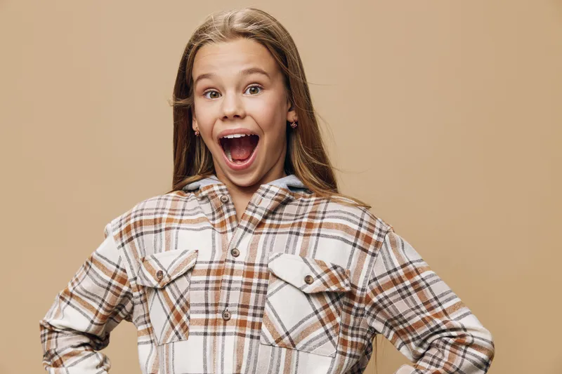Girl with long hair wearing plaid shirt showing excited expression with wide open mouth on beige background. Child standing with hands on hips for happiness and surprise emotion.