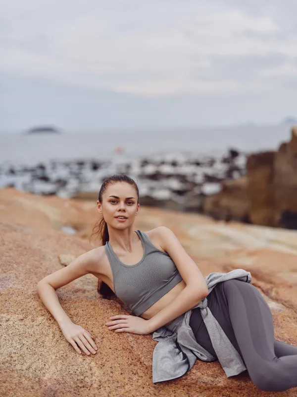 Young woman in athletic wear relaxing on rocks by the beach, showcasing fitness and tranquility in a serene coastal setting