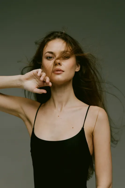 Young woman with long hair wearing black top posing confidently with hand near face against neutral background