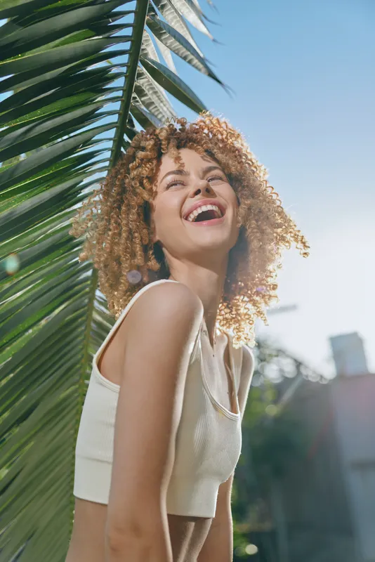 smiling woman with curly hair enjoying sunlight outdoors near green palm leaves on a bright clear day, natural joy and summer vibes