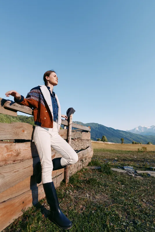 Woman leaning on wooden fence in countryside with distant mountains, wearing shearling jacket and rubber boots, gazing into clear sky enjoying peaceful outdoor leisure.