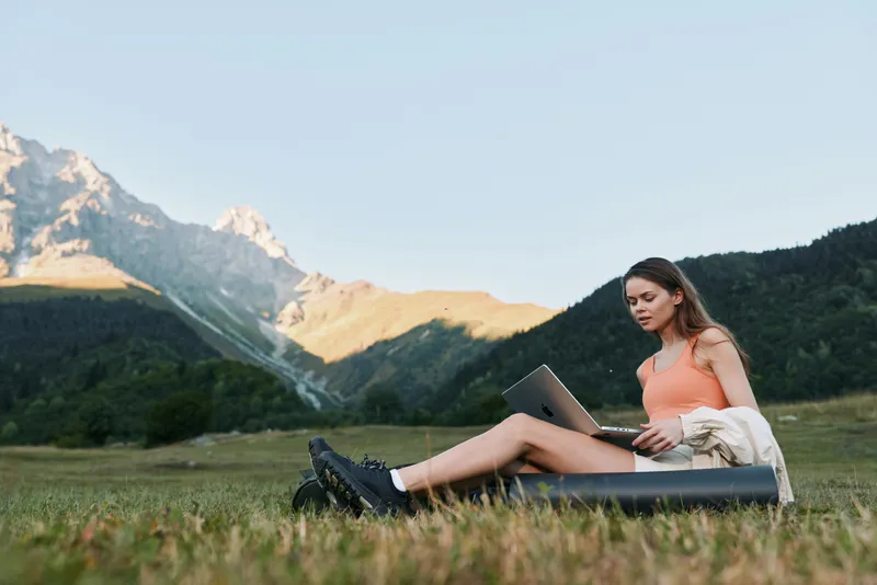 A woman sits on a grassy field with a laptop in an outdoor setting. She looks focused and relaxed, blending nature with work for a serene productivity scene.