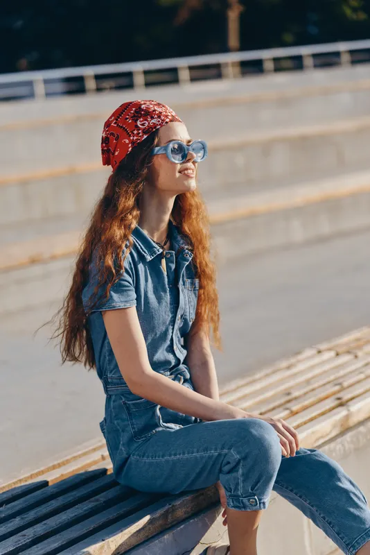 lifestyle woman in modern boho-western denim street style wearing blue denim outfit and red bandana with film color effect sitting outdoors on wooden bench