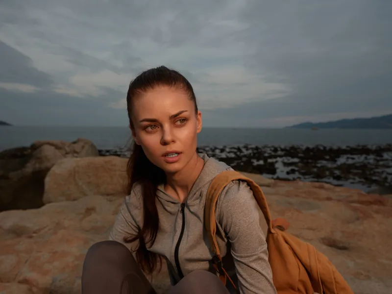 A young woman in sportswear with a backpack sits on rocky terrain by the sea, showcasing an expression of contemplation against a stunning coastal backdrop