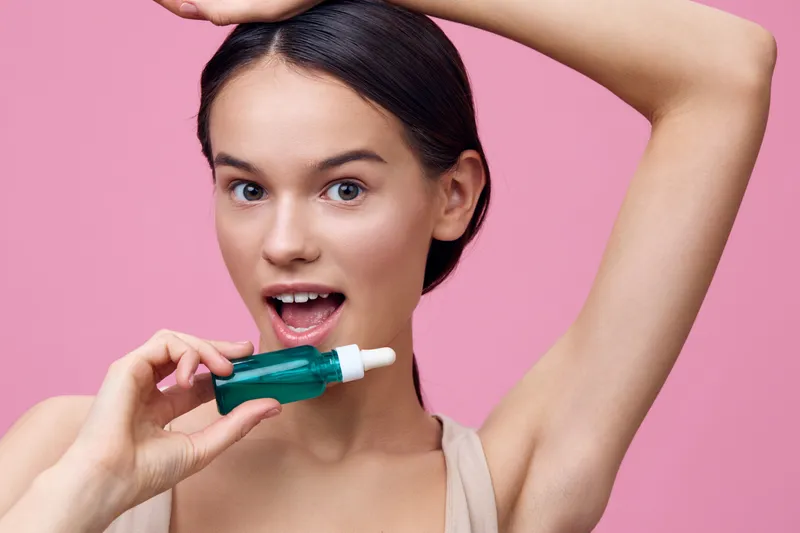 A young woman promoting a green serum for hair growth with a surprised expression against a pink background, showcasing beauty and self care
