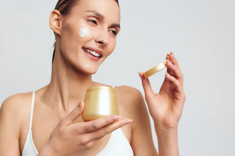 Young woman applying face cream on cheek, smiling gently, skincare routine, natural beauty, soft lighting, clean background, self care, moisturizing product, healthy skin
