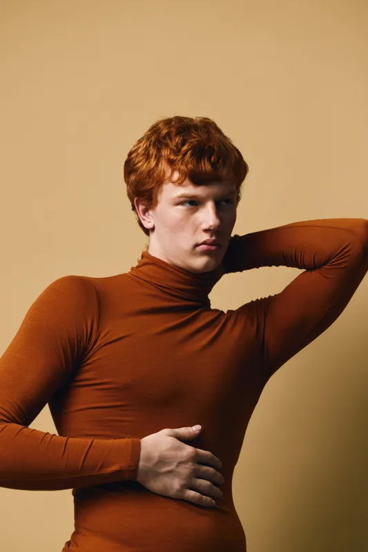 Young man with red hair and fair skin wearing brown turtleneck sweater posing with hand on stomach and arm behind head against beige background.