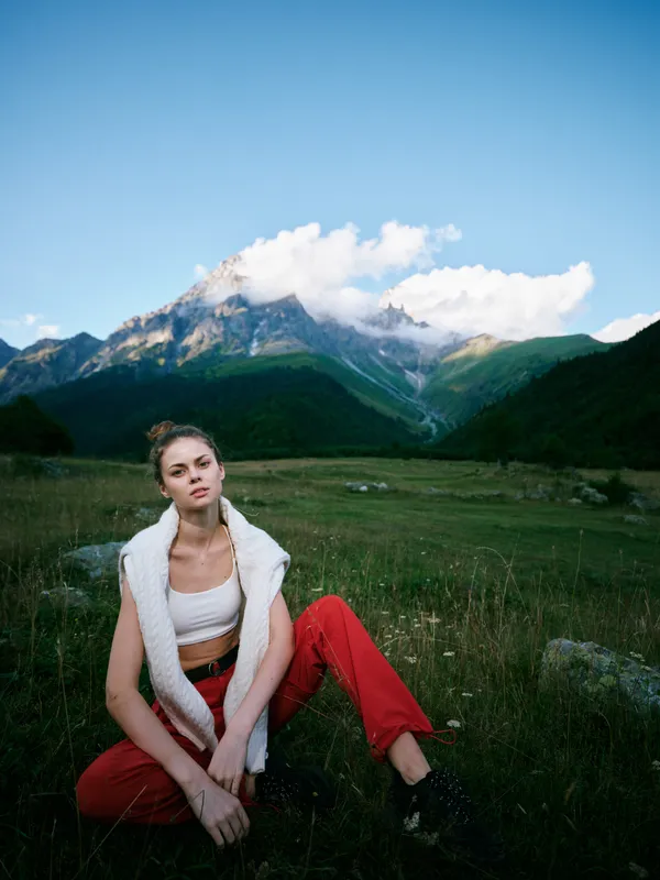 Mountain field outdoors fashion woman landscape casual scenery nature lifestyle, a relaxed pose in a meadow with distant peaks under an open sky, wind in hair, rustic tones and tranquil mood.