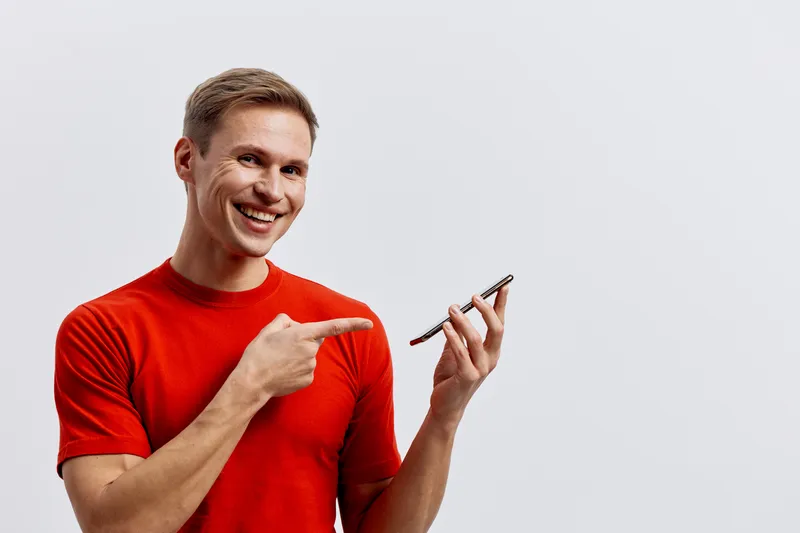 Cheerful smiling man in red t-shirt holding smartphone and pointing at it, isolated on plain white background. Casual lifestyle concept