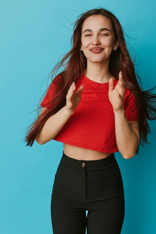 Young woman with long hair wearing red crop top and black pants clapping hands with smile on blue background. Female model with casual outfit and positive expression in studio.