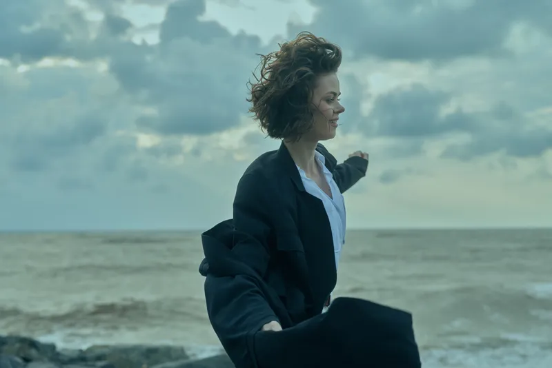 Wind sweeps across a seaside scene as a woman in a dark coat strides along the shore, capturing motion and dramatic atmosphere against cloudy skies.