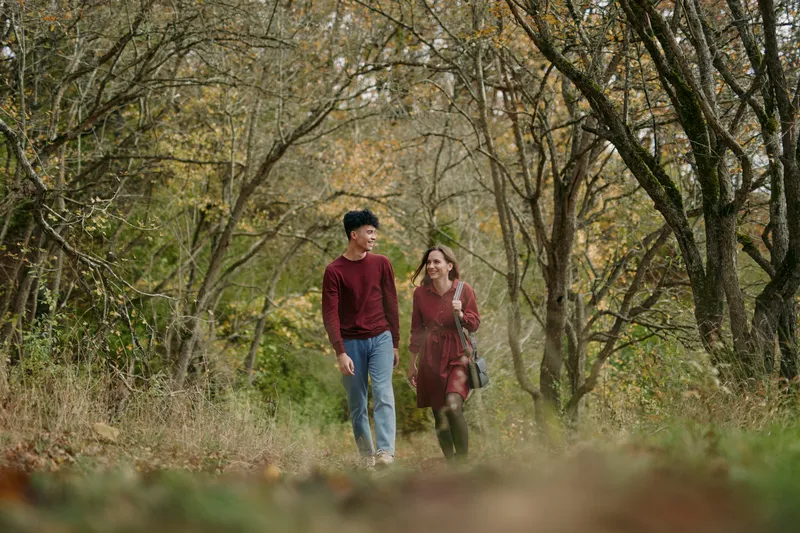 A candid, authentic scene of a couple walking along a wooded path, sharing warm smiles and natural interaction, captured in a truthful documentary style with soft autumn light.