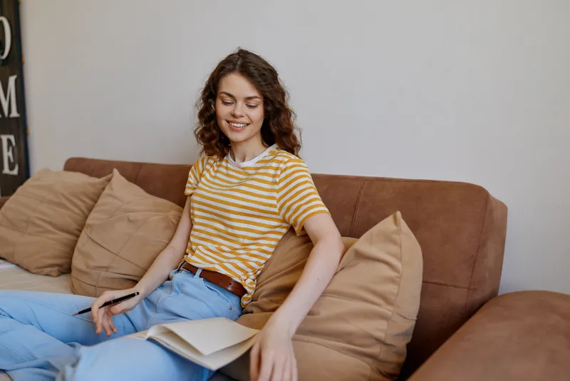 Cheerful woman indoors, relaxing on a couch, reading a book, striped tee, casual atmosphere, warm lighting, cozy living room, natural colors, candid lifestyle moment