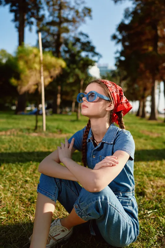 thoughtful young woman sitting on grass in park wearing red headscarf and blue sunglasses on sunny day outdoors with trees around