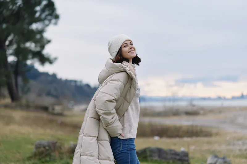 Woman in a long beige puffer coat standing outdoors by water at sunset, smiling softly, wearing a knit scarf and beanie, serene landscape, casual winter fashion for outdoor lifestyle