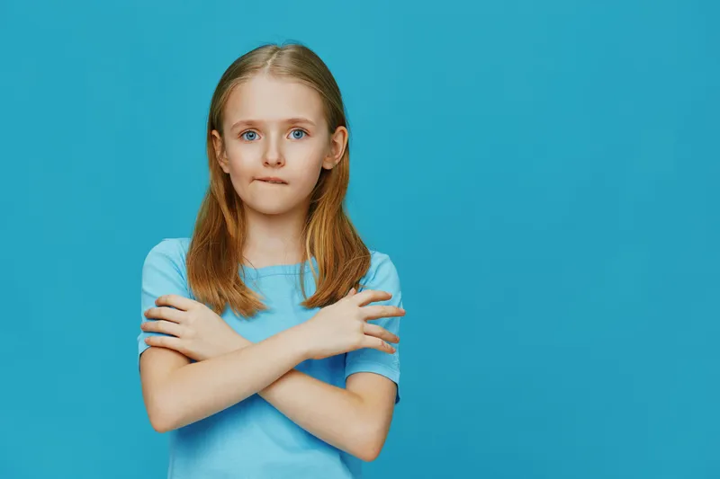A young girl with long blonde hair wearing a light blue shirt, standing with crossed arms against a bright blue background, expressing a thoughtful emotion
