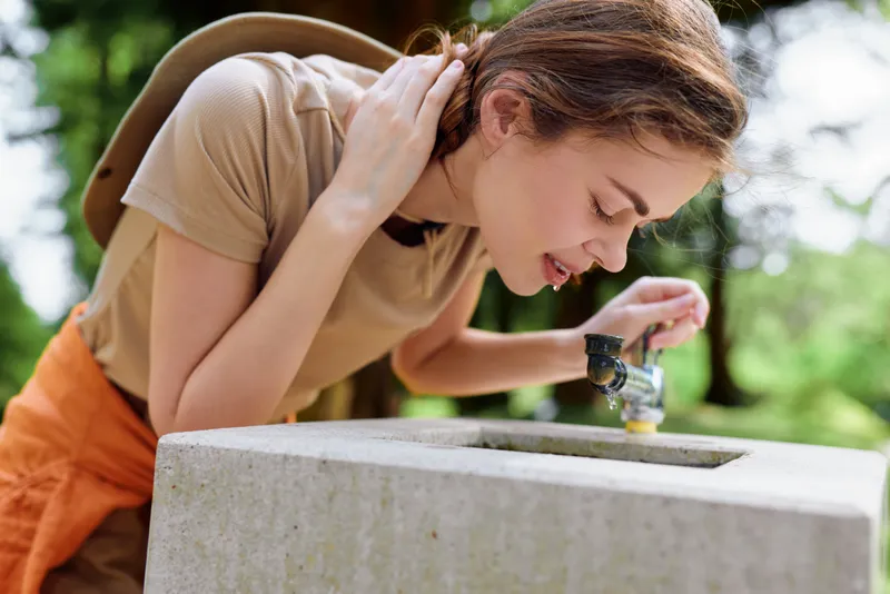 Woman drinking from a concrete fountain with a backpack in a sunny park, outdoor scene capturing hydration and summer leisure, candid portrait of casual travel and refreshment.