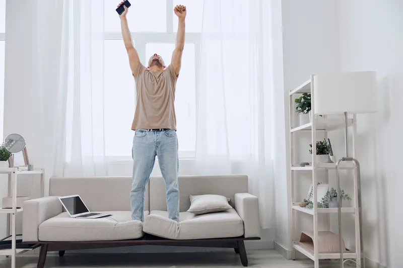 A frantically celebrating man stands on a couch, illustrating the blend of personal and work life while embracing joyful imperfection in a digital detox environment.