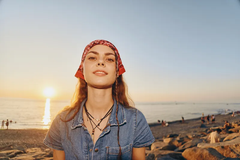 Lifestyle woman in modern boho-western denim street style stands at sunset on rocky beach with film color tone, expressing relaxed confidence and free spirit vibe.