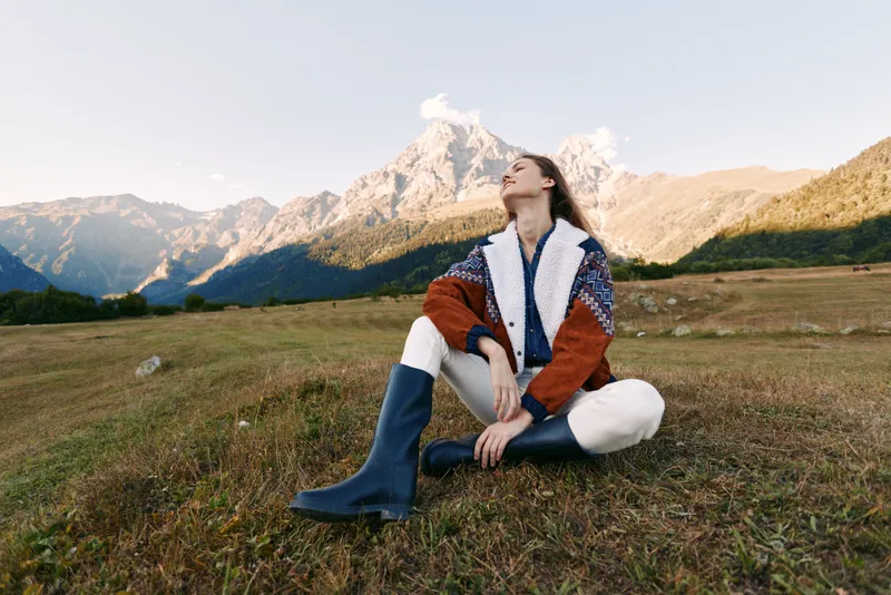 Woman mountains meadow boots sweater travel hiking, relaxed portrait of a young woman sitting in a grassy alpine meadow wearing knit sweater and boots, enjoying scenic mountain landscape.