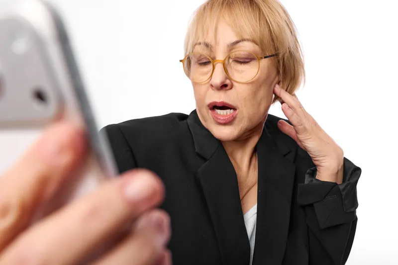 Elderly woman in business suit and glasses shows anxiety and stress while holding her ear during a phone call, worried face, blurred mobile in foreground, communication concept.
