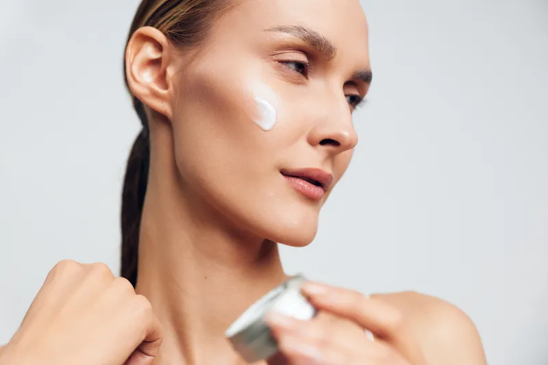 Close-up of a young woman with fair skin applying white cream to her cheek in a soft studio portrait, calm mood, neutral background, skincare concept.