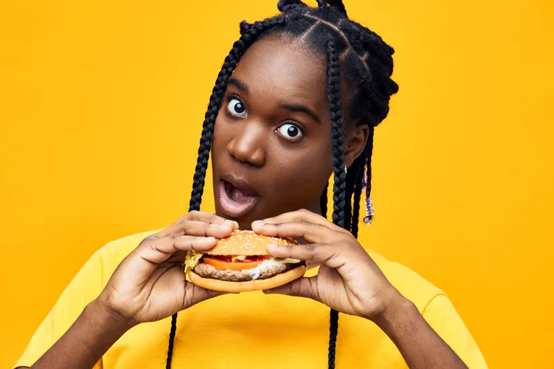 Surprised young woman of African descent holding a delicious burger, wearing a bright yellow t shirt, set against a vibrant yellow background